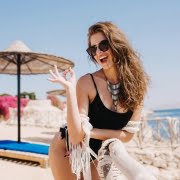 Shapely cheerful girl in black bodysuit laughing and posing with okay sign on the sea beach. Attractive young lady in trendy necklace cute smiling and having fun on resort in summer weekend..